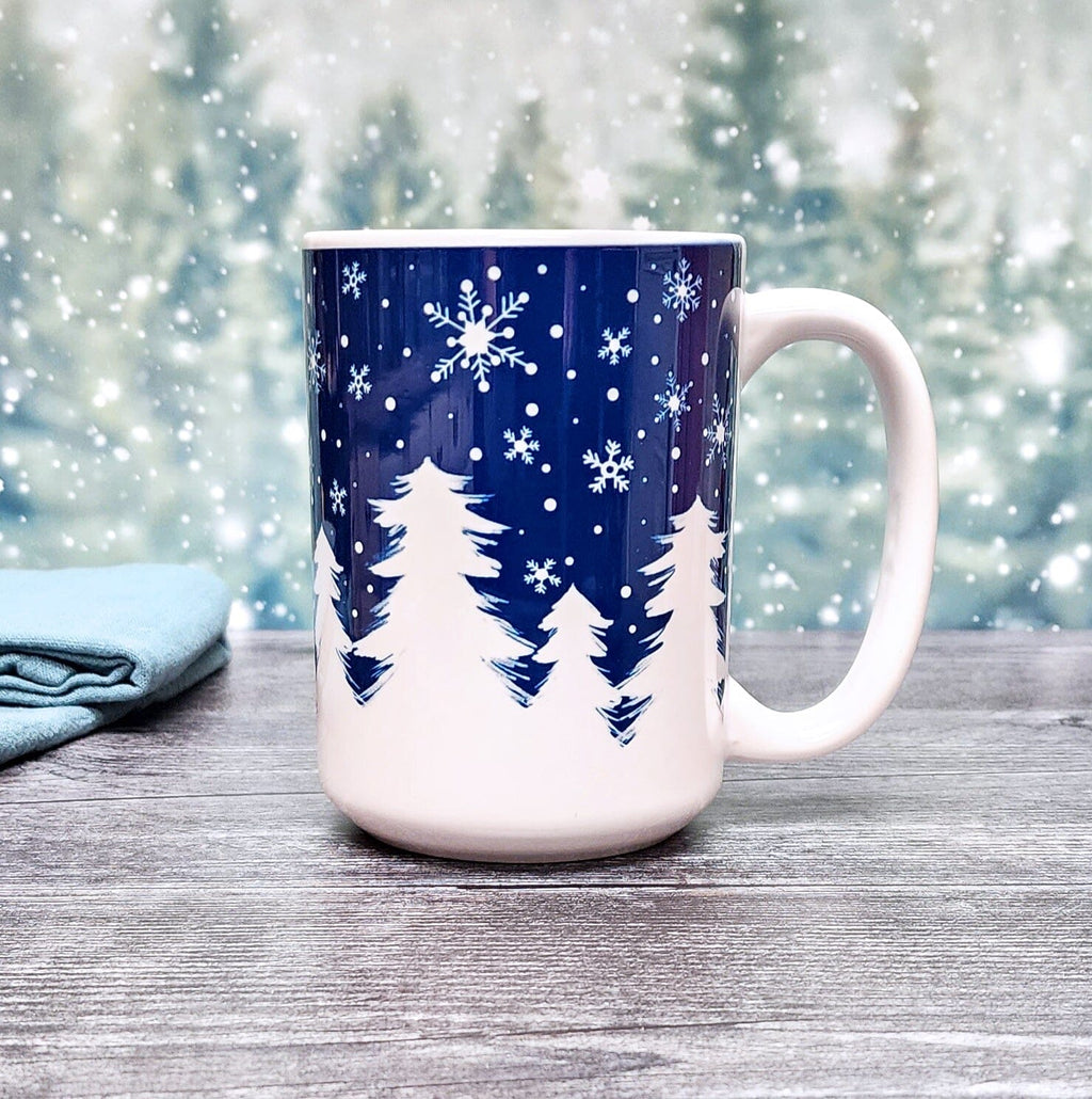 Winter Night Snow Mug (15oz) on a festive snowy tree background, from Amy's Coffee Mugs. A ceramic coffee mug designed with an illustrated winter night snow treeline. The design features a navy blue sky filled with white snowflakes over a row of white pine trees along the bottom.